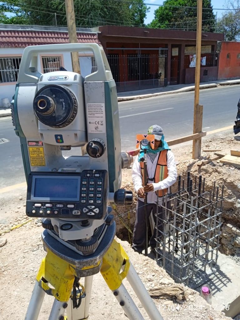 Primer plano del panel de control de una estación total topográfica en una vialidad urbana, con un topógrafo sosteniendo un prisma de medición al fondo.