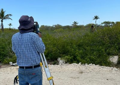 Imagen: La foto de la izquierda, con el topógrafo de espaldas y vegetación densa. Texto Alternativo (Alt Text): Un topógrafo profesional con sombrero de ala ancha opera una estación total en trípode en un terreno exterior rocoso. Detrás hay vegetación densa de matorrales y palmeras bajo un cielo azul despejado.