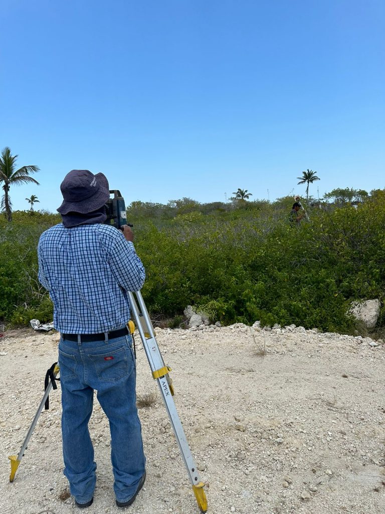 Imagen: La foto de la izquierda, con el topógrafo de espaldas y vegetación densa. Texto Alternativo (Alt Text): Un topógrafo profesional con sombrero de ala ancha opera una estación total en trípode en un terreno exterior rocoso. Detrás hay vegetación densa de matorrales y palmeras bajo un cielo azul despejado.