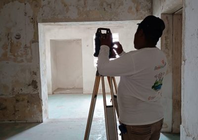 Un topógrafo profesional con camisa blanca de manga larga con logotipos (como el de Geoland) y gorra opera una estación total en trípode en el interior de una habitación con paredes de piedra y yeso rústicas y desgastadas.