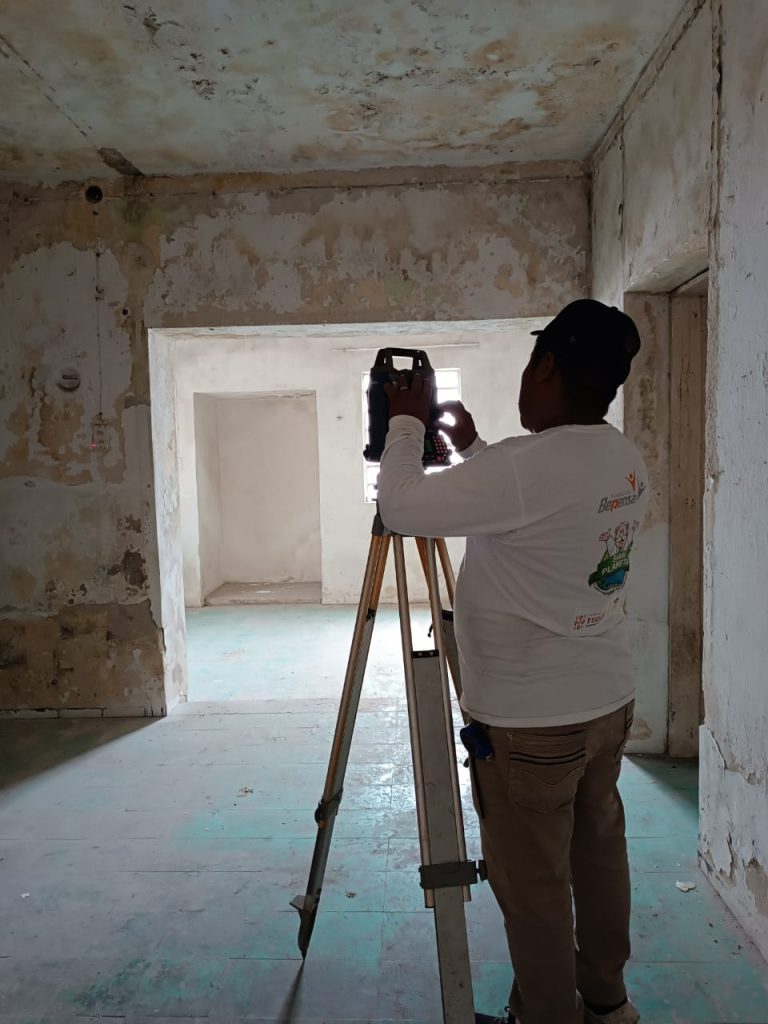 Un topógrafo profesional con camisa blanca de manga larga con logotipos (como el de Geoland) y gorra opera una estación total en trípode en el interior de una habitación con paredes de piedra y yeso rústicas y desgastadas.