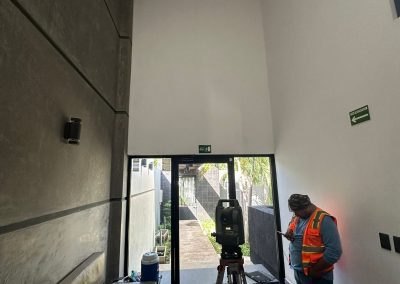 Un topógrafo con chaleco reflectante naranja y gorra manipula una estación total en trípode dentro de un pasillo interior estrecho de un edificio moderno con paredes de hormigón texturizado y señalización de emergencia verde. Hay una vista exterior al fondo.