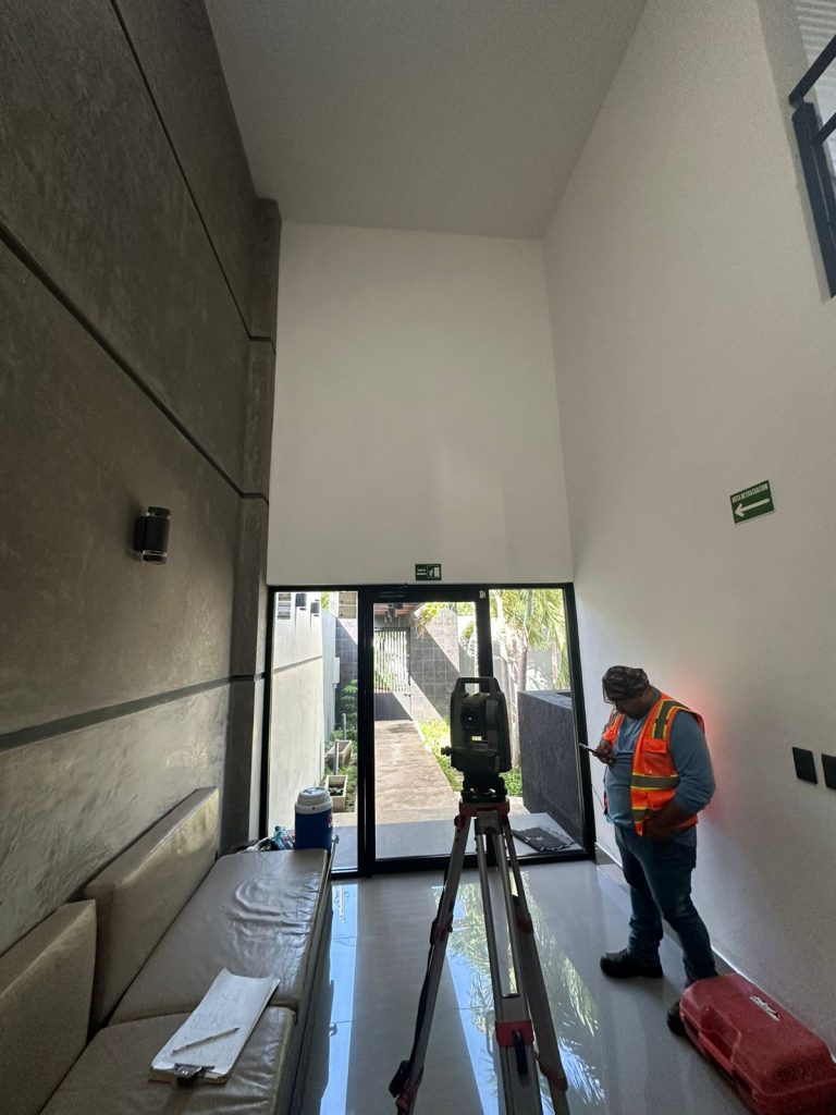 Un topógrafo con chaleco reflectante naranja y gorra manipula una estación total en trípode dentro de un pasillo interior estrecho de un edificio moderno con paredes de hormigón texturizado y señalización de emergencia verde. Hay una vista exterior al fondo.