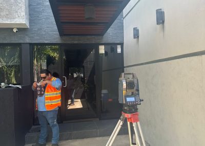 Un topógrafo con chaleco reflectante naranja y gorra ajusta una estación total en trípode frente a la entrada de vidrio de un edificio de diseño moderno con detalles arquitectónicos de madera y paredes de paneles grises.