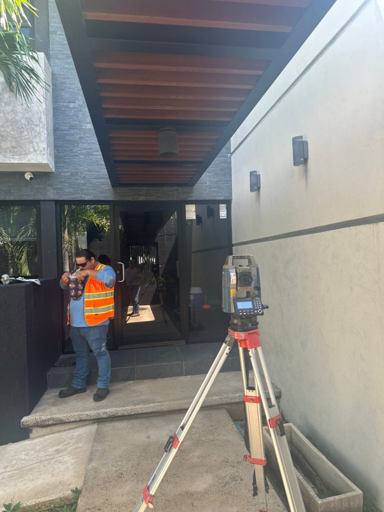 Un topógrafo con chaleco reflectante naranja y gorra ajusta una estación total en trípode frente a la entrada de vidrio de un edificio de diseño moderno con detalles arquitectónicos de madera y paredes de paneles grises.